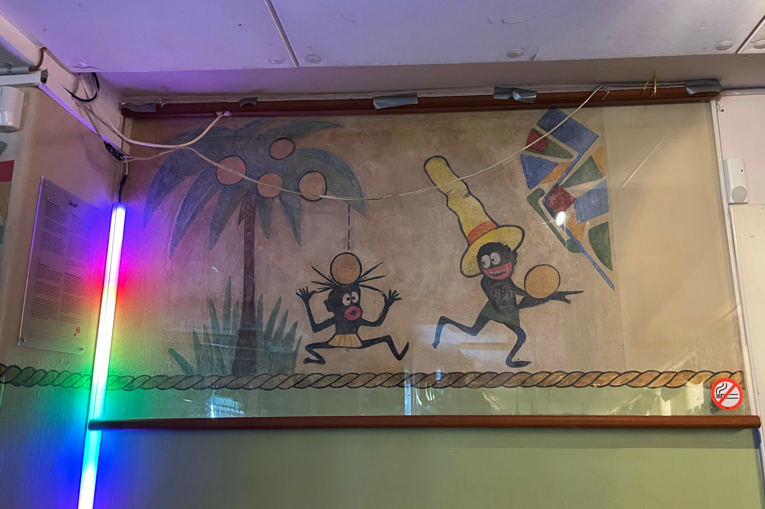 Ved siden af vægmaleriet er en plade med information om Storm P ledsaget af følgende tekst på både dansk og engelsk: »Helt typisk for den tid i begyndelsen af forrige århundrede var et vestligt, chauvinistisk syn på udlændinge og etniske minoriteter. Racemæssige kendetegn blev trukket frem og udstillede – set med nutidens øjne – tidens markante fordomme og uvidenhed om andre kulturer hos både tegnere og publikum. Mens tegningerne dengang virkede både harmløse og sjove, kan de i dag virke både stødende og uacceptable. 
Storm P. udførte en lang række vægudsmykninger i stil med udsmykningen her. I dag findes der kun tre bevarede vægmalerier af Storm P.. Det, der er tilbage på malerierne på A-vej, blev i sommeren 2022 restaureret af Nationalmuseet. Karikaturerne, der kan vække anstød, er således bevaret. Dels af hensyn til den historiske kulturarv, dels som en nutidig mulighed for refleksion over, at vores syn på mennesker og kulturer og bevidsthed om diversitet, inklusion og diskrimination i dag er et andet sted end for 100 år siden, da Storm P Slap sine karikaturer løs.«