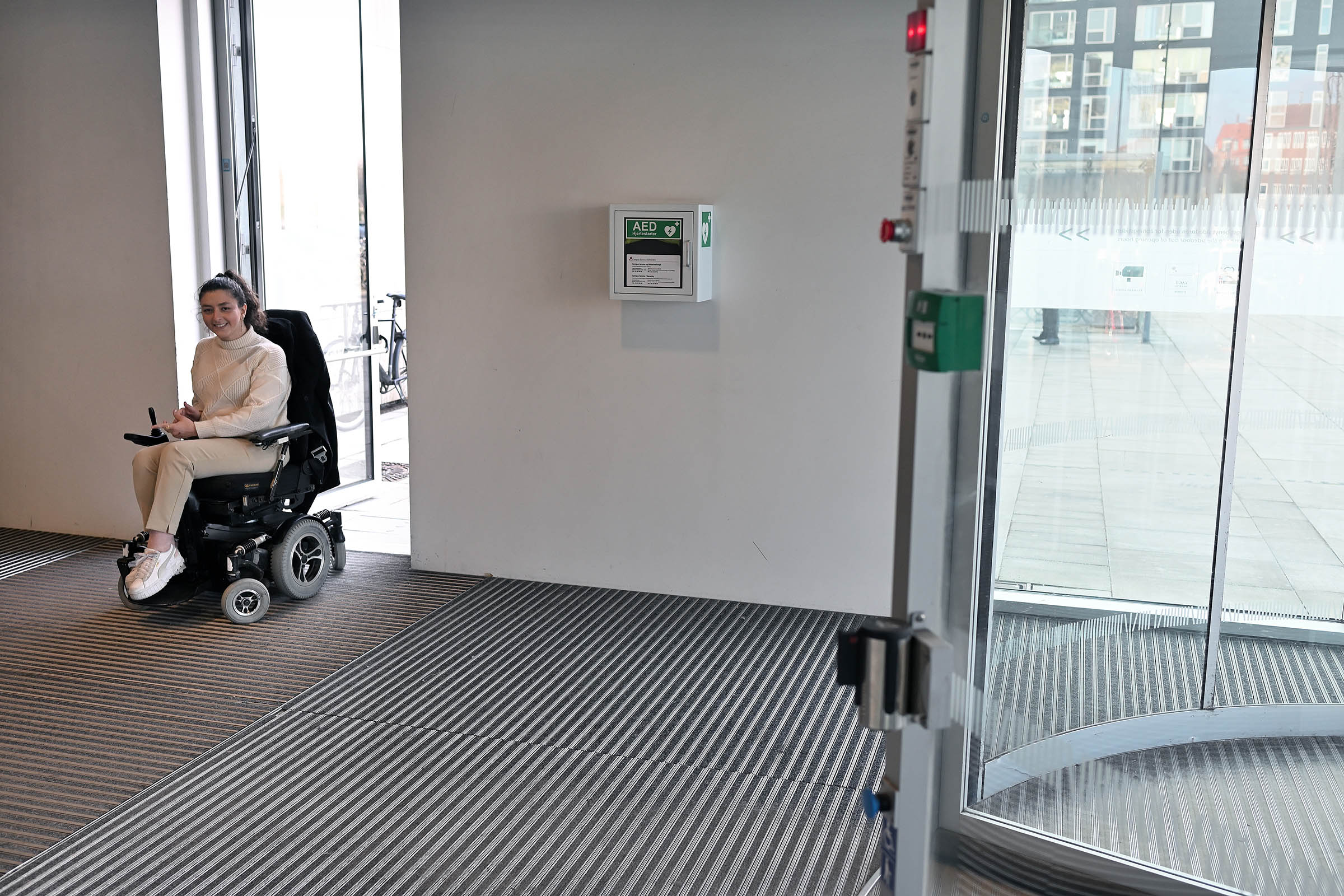 Emilie Ghali triller ind ad fakultetets hovedindgang. Døren åbner automatisk, og der er intet trin, så den elektriske kørestol kommer nemt igennem.