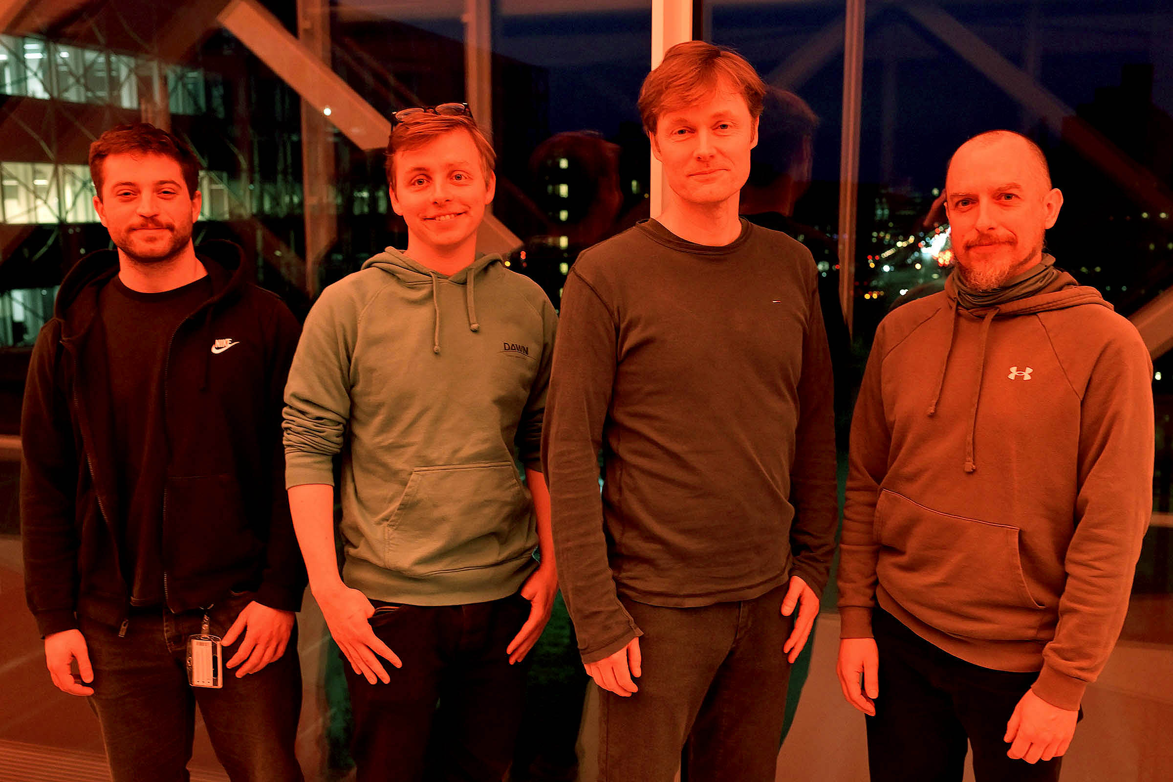 Fire fysikere i rødt lys i Niels Bohr Bygningens gangbro over Jagtvej. Fra venstre er det Georgios Nikopoulos, Albert Sneppen, Darach Watson og Gabriel Brammer. 